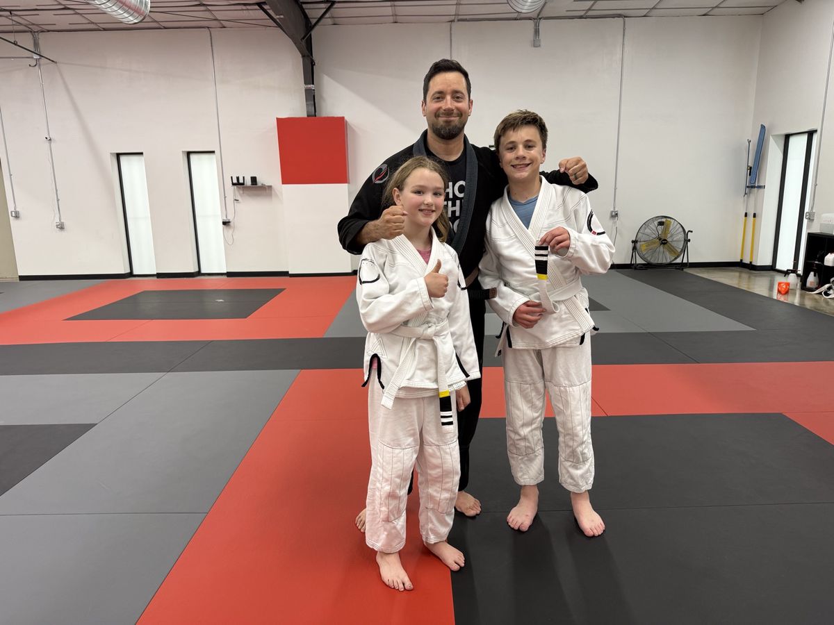 Coach Alex with two younger Journey students in white gis giving thumbs up on the mat