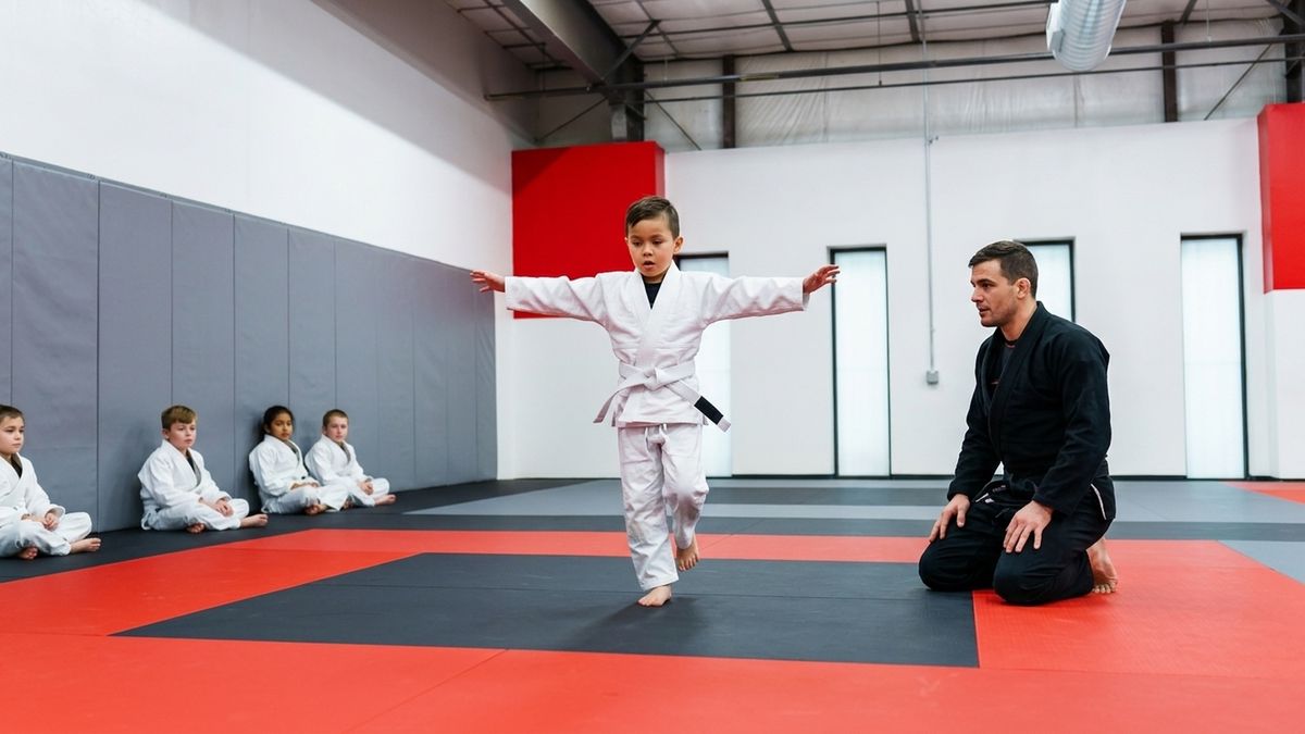 A child in a white gi standing on one foot on a mat, arms out for balance, coach kneeling nearby watching