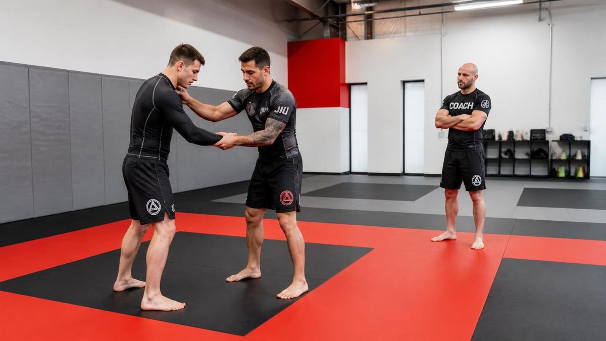 two training partners drilling a standing grip technique in a bright gym, one clearly newer and being guided, coach observing nearby