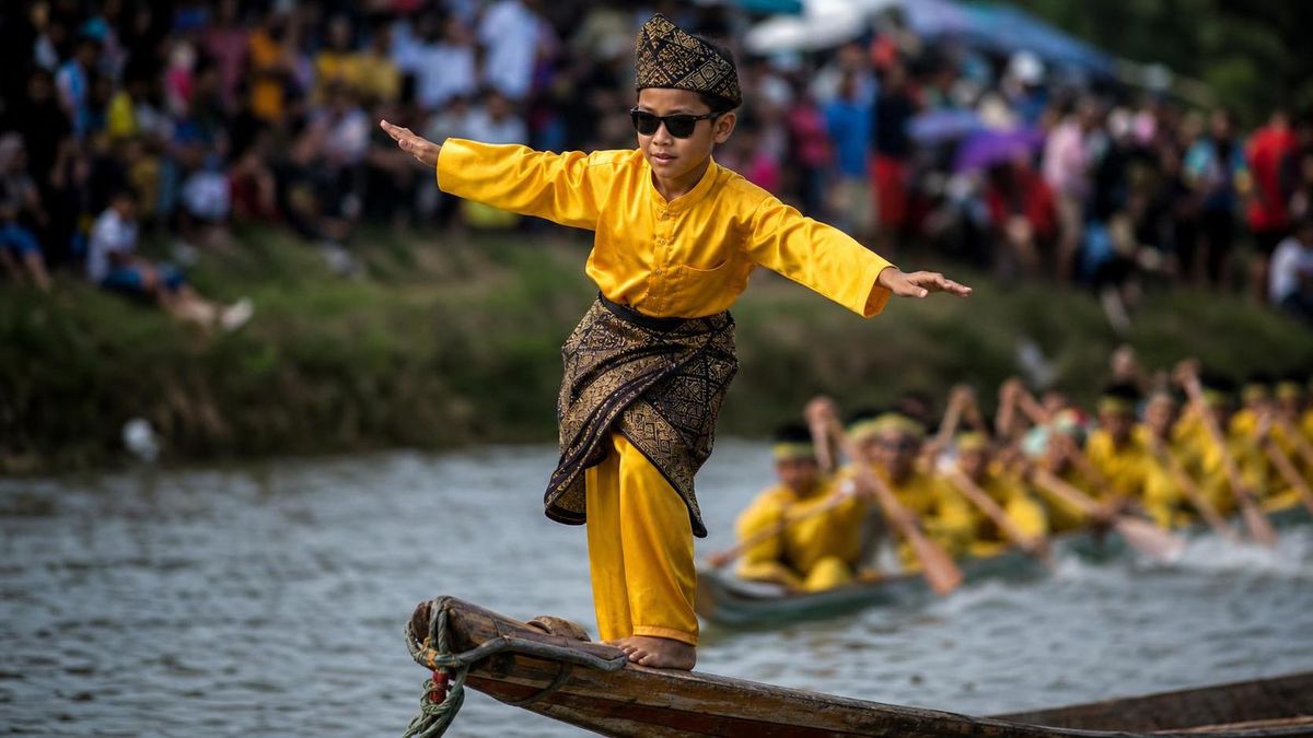 A young boy in traditional Indonesian dress and dark sunglasses balancing on the bow of a long racing boat, arms out, crowd blurred behind him.