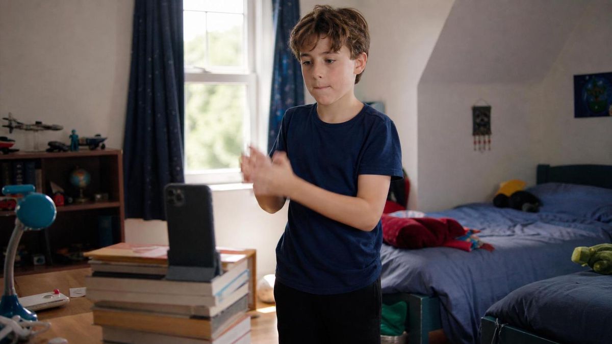 A kid in a bright ordinary bedroom re-recording the same short phone video for the fifth time, phone propped on a stack of books, caught mid-reset between takes with a flat neutral expression, natural daylight.