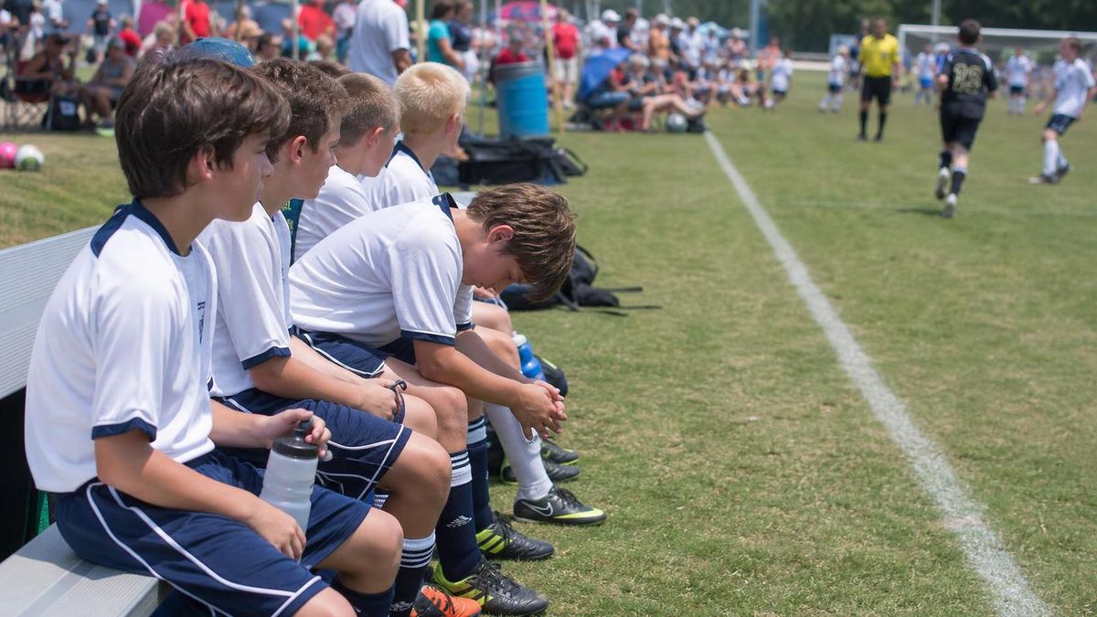 a row of kids on a sideline bench in soccer gear, one of them staring down at his cleats while the game goes on behind him