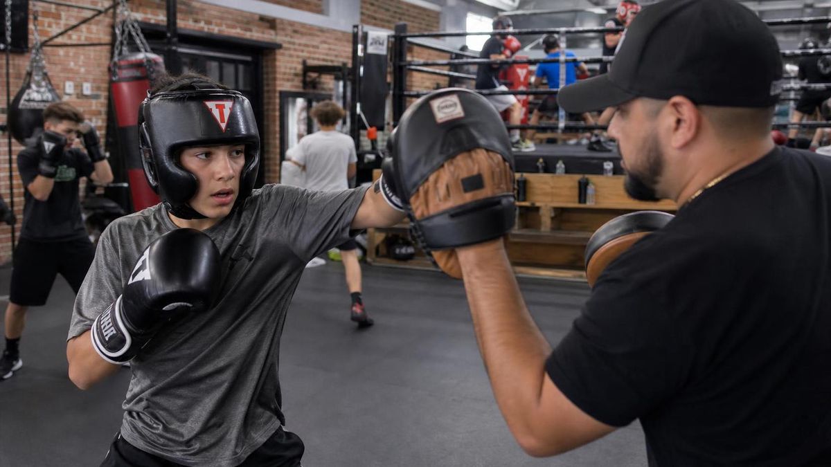 a teenage boy in boxing gloves and headgear working the mitts with a coach in a small gym, other kids training in the background