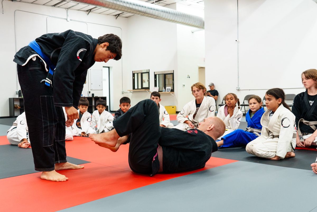 A Journey BJJ instructor demonstrating a technique to seated kids in white gis