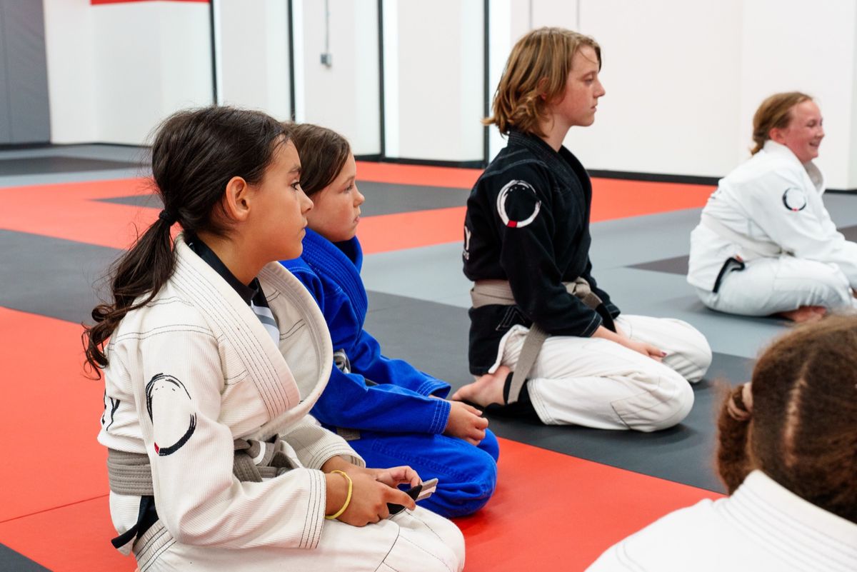 Kids in white gis seated and attentive on the Journey BJJ mats