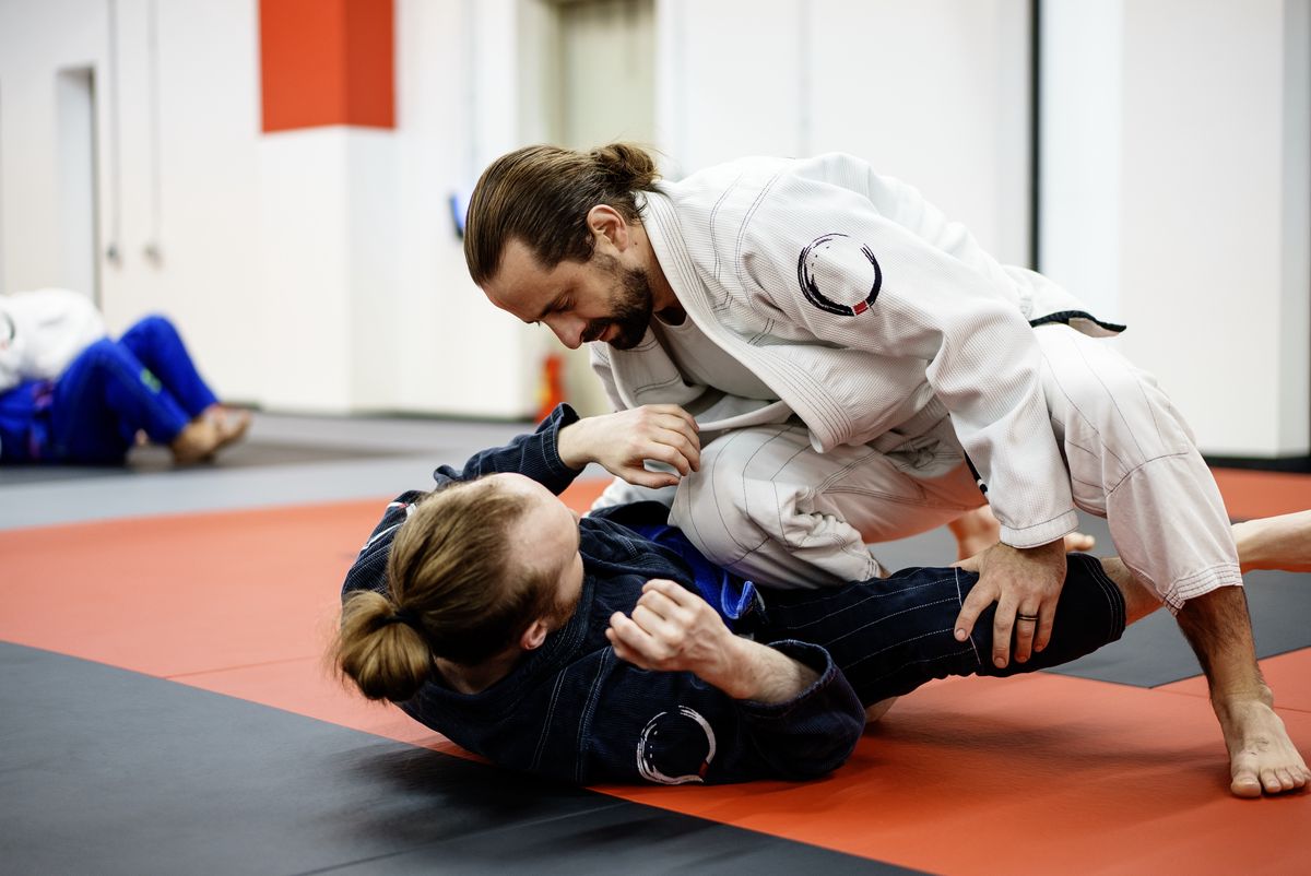 Two adult students in gis doing live positional sparring on the mat, one working to keep base while the other off-balances them (real gym photo)