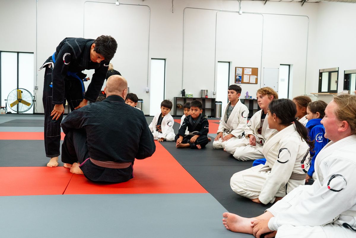 A Journey BJJ kids class seated in a row on the mat watching a coach demonstrate a technique with a partner, wide framing showing the full room.