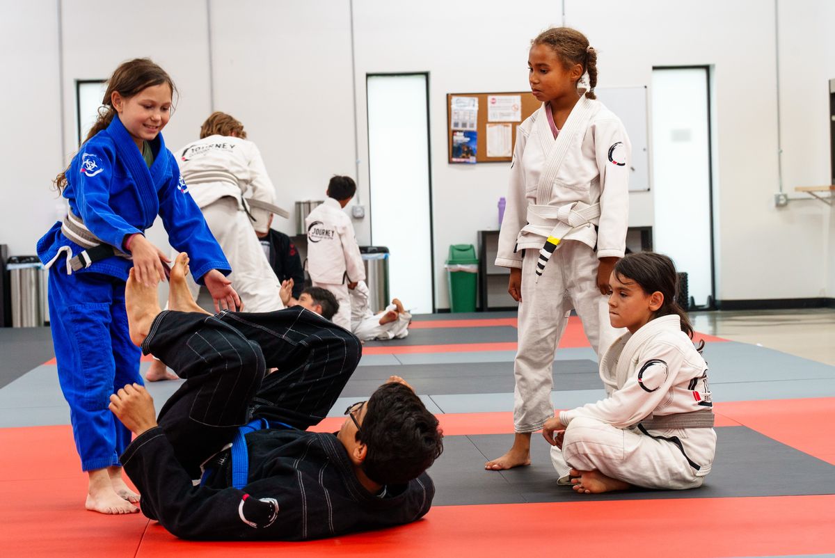 a girl in a white gi working a technique with a partner on the mat while two other kids watch from beside them
