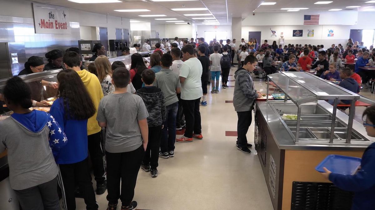 a busy school cafeteria at lunch, a crowded hot-food counter with students packed in line while the salad and vegetable station beside it sits untouched