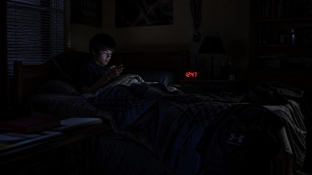 A dark bedroom at night, alarm clock reading 12:47 AM, a teenager still awake sitting up in bed.