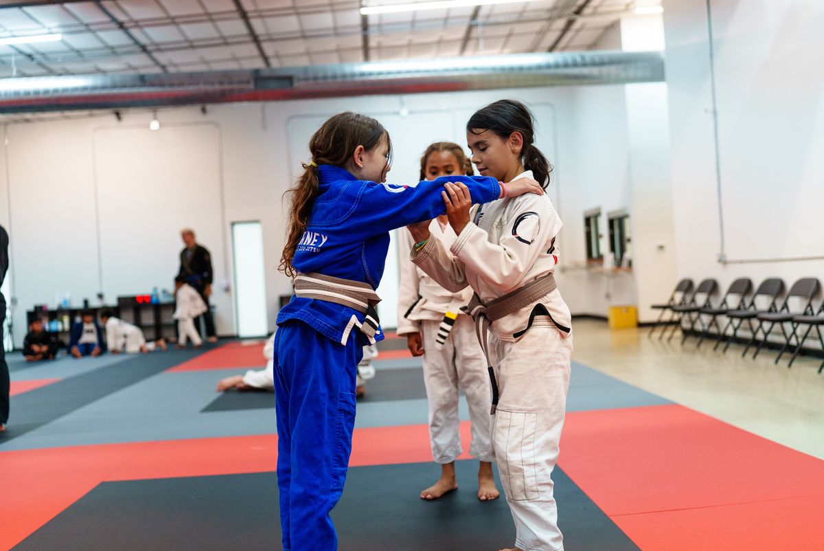 Two kids standing in a Madison gym gripping each other's gi sleeves mid grip-fight, both upright on their feet, coach watching