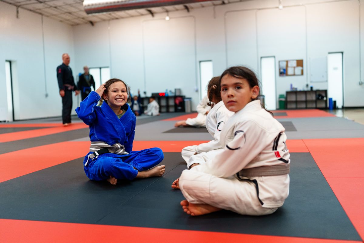 REAL GYM PHOTO NEEDED (no AI): two kids in gis kneeling upright on the mat between drills, laughing, one helping the other fix a belt