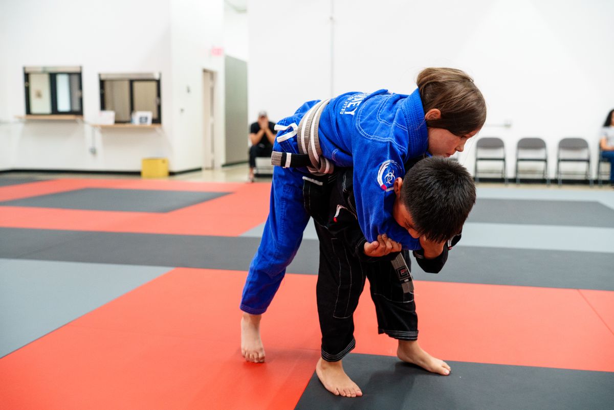 Two kids in gis on the mat mid-drill in a bright gym, both concentrating hard, one guiding the other's grip.